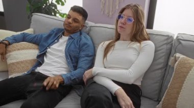 Modern bir oturma odasındaki koltukta dinlenen bir çift, uzaktan kumandalı bir adam, gözlüklü bir kadın, yastıklar ve bitkilerle çevrili, kapalı bir apartman dairesinin keyfini çıkarıyor..