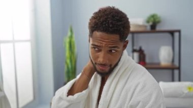 Beyaz cüppeli yakışıklı adam arka planda bitki ve raflarla dolu aydınlık bir odada otururken gülümsüyor.