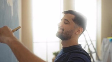 Evde, oturma odasında resim yapan genç bir adam. Parlak doğal ışık ile bir yenileme projesi üzerinde çalışırken gülümsüyor..