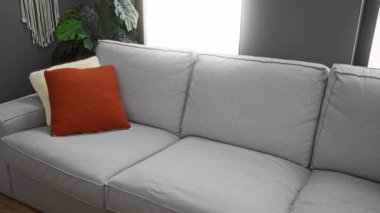 Genç adam gri bir koltukta oturuyor modern bir oturma odasında, üzgün ve üzgün görünüyor, beyaz polo bir gömlek giyiyor, arkasında kırmızı bir yastık ve yeşil bitkiler.