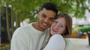 Şehir parkında kucaklaşan gülümseyen çift arka planda yeşillikle olan mutlu ilişkilerini sergiliyor rahat ve sevgi dolu bir atmosfer yaratıyorlar..