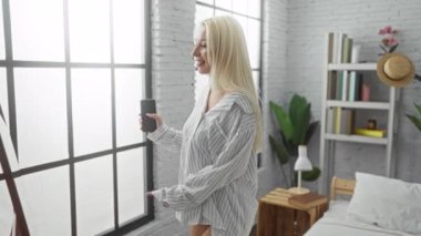 Sarışın kadın yatak odasında elinde telefon tutarak mutlu bir şekilde dans ediyor. Etrafı geniş pencereli, kitaplı ve bitkilerle çevrili. Rahatlatıcı atmosferi vurguluyor..