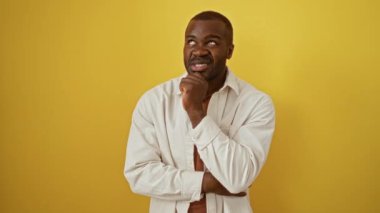 Dostum, Afro-Amerikan adam tişört giyiyor, endişeli ve gergin, elleri çenesinde, izole edilmiş sarı arka planda, endişeli ve bir soru hakkında düşünüyor