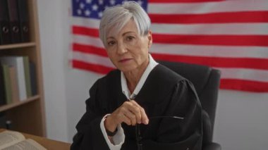 Arka planda Amerikan bayrağı olan yaşlı bir kadın yargıç, elinde gözlük tutarak Birleşik Devletler mahkemesinde ciddi ve düşünceli bir tavır takınıyor..