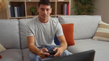 Rahat bir oturma odasında oynayan genç bir adam modern dekorla çevrili neşeli bir ifadeyle bir denetleyiciyi tutuyor, rahat ve eğlenceli bir ev atmosferini vurguluyor.