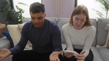 Sıcak bir oturma odasında birlikte oturan bir çift, kadın gözyaşlarını silerken, erkek el kol hareketleri yapıyor, bir ilişki içinde duygusal sohbet ve destek öneriyor.