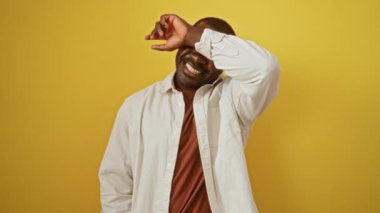 Afro-Amerikalı bir adam, tişörtünü giymiş neşeli bir şekilde dikiliyor, dikizliyor, gülümsüyor ve izole edilmiş sarı arka planda ellerini gösteriyor.