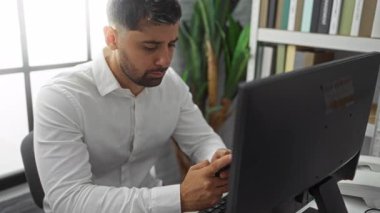 Ofiste akıllı telefon kullanan genç bir adam masasında otururken bitki ve kitaplıkla, işine odaklanmış..