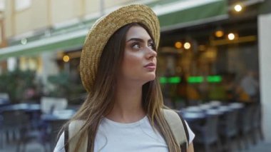 Hasır şapkalı bir kadın şehir ortamında bir kahve dükkanının önünde geziyor, şık bir moda sergiliyor ve kadın güzelliğine ve şehir atmosferindeki açık hava cazibesine odaklanmış rahat bir hava veriyor..