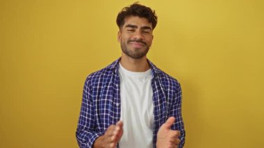 Gülümseyen Latin asıllı, günlük ekose gömlekli genç adam canlı sarı bir arka plana el çırpıyor, özgüven ve orjinallik yayıyor..