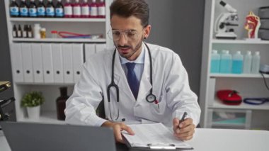 Doktor önlüğü ve gözlüğü giyen genç bir adam hastane odasında, tıbbi ekipman ve dosyalarla çevrili, profesyonel bir sağlık hizmeti sunan notlar yazıyor..