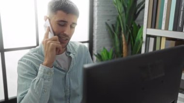 Ofis ortamında telefonla konuşan, bilgisayarla masaya oturan, günlük gömlekler giyen, ev bitkileri ve kitaplarla çevrili, modern bir iş ortamını yansıtan genç bir adam..