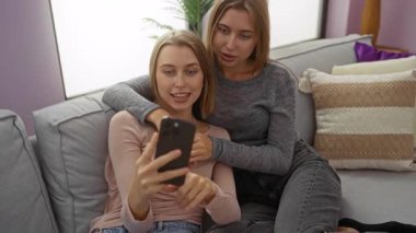 Kız kardeşler rahat bir oturma odasında neşeli bir video çağrısı yapıyorlar, aile bağlarını ve dijital etkileşimlerini gösteriyorlar, gülümsemeler ve rahat ev atmosferi ile sıcak ev ortamını güçlendiriyorlar..