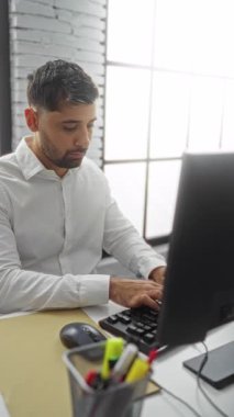 Beyaz gömlekli genç adam, modern ofis ortamında, bilgisayarlı bir masada otururken, parlak ve çağdaş bir çalışma alanında pozitifliğini dile getiriyor..