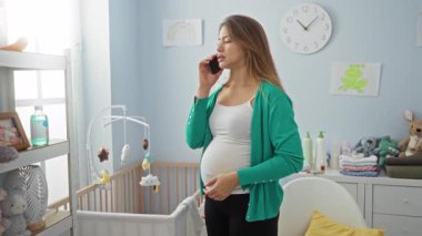 Yatak odasında hamile bir kadın beşiğin yanında durup telefonla konuşuyor. Bebek malzemeleri ve yumuşak dekorla çevrili samimi bir iç dekorun görüntülerini yakalıyor..