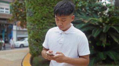 Beyaz gömlekli genç adam şehir parkında akıllı telefon kullanıyor. Arka planda yeşil ve şehir caddeleri var. Nişanlı ve dışarıda mutlu görünüyor..