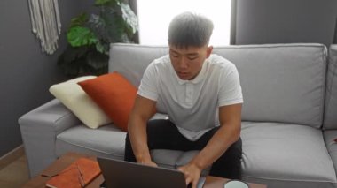 Genç adam evdeki kanepede dizüstü bilgisayarla dinleniyor. Arka planda yastıklar ve bitkilerle rahat bir oturma odasında yorgun görünüyor..