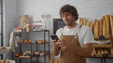 Fırının içinde akıllı telefon kullanan genç bir adam önlük takıyor, arka planda çeşitli ekmek ve hamur işleri var, odaklanmış ve meşgul görünüyor.