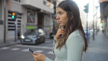Elinde telefon tutan genç bir kadın şehir caddesinde duruyor. Şehir trafiği ve binaların ortasında güpegündüz düşünceli görünüyor..