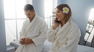 Spa elbiseli bir çift, telefonda gülümseyen bir kadın ve içeride rahatlama ve bağlantı sağlayan parlak bir sağlık odasında akıllı telefon kullanan bir erkek..