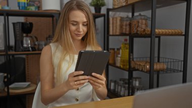 Modern ofis ortamında çalışan, tabletle meşgul, çeşitli mutfak malzemelerini gösteren raflarla çevrili, üretken ve sıradan çalışma ortamını yansıtan bir kadın..