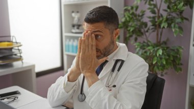 Klinikte steteskop takmış, düşünceli ve odaklanmış görünen İspanyol bir adam, iş yerinde bir tıp uzmanını öneriyor..