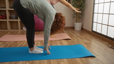 Spor salonundaki paspaslarda yoga yapan iki orta yaşlı kadın, ahşap zemin ve yoga ekipmanlarıyla dolu parlak, ferah bir odada dostluk ve fitness vurguluyor..