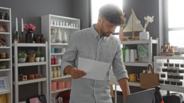 Dekor dükkanındaki genç adam çeşitli dekoratif eşyalar, bitkiler ve raflarla çevrili bir şekilde bir kağıda bakıyor..