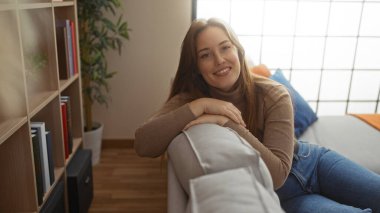 Rahat bir oturma odasında rahatlayan bir kadın, gülümseyen ve kanepede yaslanan, kitaplar ve bitkilerle çevrili, ev içinde huzurlu bir yaşam tarzını ön plana çıkaran bir kadın..