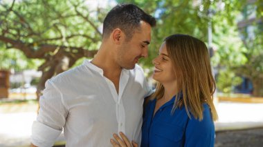Güneşli bir şehir parkında gülümseyen bir çift, bir ağacın altında kucaklaşan bir adam ve kadın, şehir ortamında sevgi ve mutluluk gösteriyorlar..
