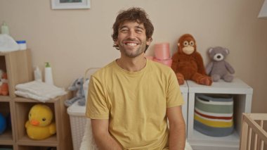 Young hispanic man smiling in a cozy bedroom with a cradle in the background, surrounded by soft plush toys, creating a warm, inviting home interior with family vibes. clipart