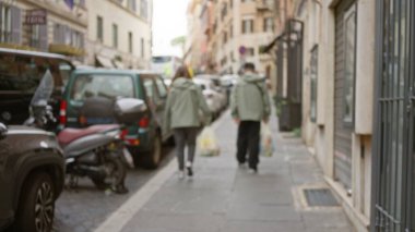 Roma, İtalya 'da eski bir caddede yürüyen, Bokeh etkisindeki araba ve binalarla günlük hayatı yakalayan iki kişinin görüntüsü bulanık..