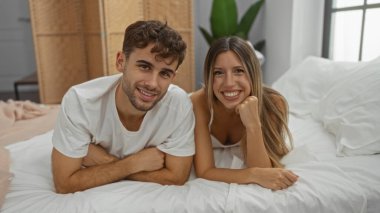 Çift, rahat bir yatak odası ortamında birlikte gülümsüyor. Kadın ve erkek rahat bir yatakta uzanıyorlar. Parlak bir evde sevgi ve mutluluk sergiliyorlar..