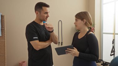 Spor salonunda elinde tablet tutan kadın, erkeklerle sohbet ediyor, spor salonu ortamında spor, teknoloji ve etkileşim sergiliyor, takım çalışmasını ve eğitim atmosferini vurguluyor..