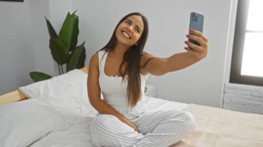 Kadın rahat bir yatak odasında selfie çekerken gülümsüyor, beyaz çarşaflı bir yatakta oturuyor ve arka planda bir saksı bitkisi, rahat ve neşeli bir atmosfer taşıyor..