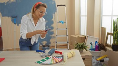 Yeni dekore edilmiş oturma odasında renk örneklerini fotoğraflayan bir kadın, parlak bir öğleden sonra evi yenileme ve boyama araçlarını sergiliyor..