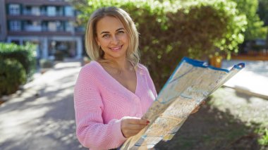 Güneşli bir günde haritayla şehir caddesini keşfeden, pembe kazak giyen, sarı saçlı, açık havada duran, şehir manzarası ile çevrili, seyahat macerasından zevk alan bir kadın..