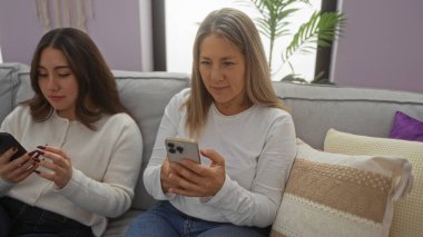 Anne ve kızı rahat bir oturma odasındaki kanepede telefon kullanıyor, aile bağlarını ve modern teknolojiyi ön plana çıkarıyor..