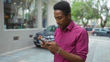 Şehirdeki genç adam akıllı telefon kullanıyor, günlük gömlek giyiyor, arka planda arabalar olan ekrana odaklanmış, modern bir şehir havası yakalıyor..