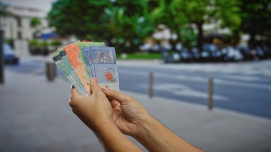 Bir şehir caddesinde elinde Malezya sirkesi banknotları tutan adam yeşil ağaçlı ve binalı bir şehir zeminine karşı çeşitli para birimleri ve iş konsepti sergiliyor..