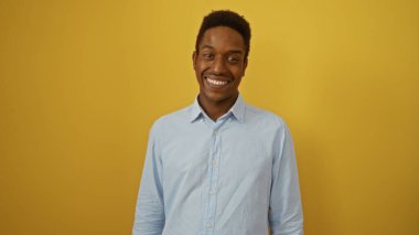 İzole edilmiş sarı arka plana karşı güvenle gülümseyen genç yakışıklı bir adam. Günlük mavi gömlek giyiyor..