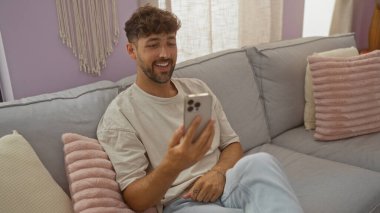 Oturma odasındaki bir kanepede rahat bir şekilde oturan genç adam telefonuna bakıp gülümsüyor.