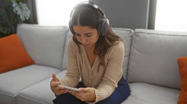 Rahat oturma odasında akıllı telefon ve kulaklık kullanan, yumuşak kanepeli ve eğlence için sıcak ışıklandırmalı bir kadın..