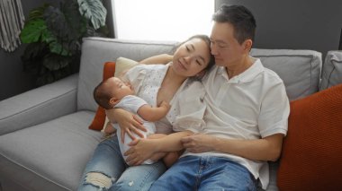 Aile oturma odasında kanepede dinlenirken, anne kucağında bebek, baba ise sıcak bir yuva kurarken, sevgi ve beraberlik sergiliyor..