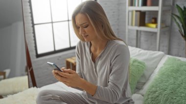 Sıcak yatak odasında, yeşil yastıkları ve kitaplığı olan akıllı telefonu kullanarak yatakta otururken kaşlarını çatan kadın, gündüzleri evde tefekkür ve rahatlığı simgeliyor..