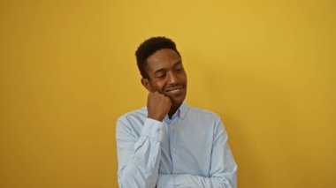 İzole edilmiş sarı bir duvara güvenle gülümseyen genç bir adam modern bir tarz sergiliyor, yakışıklı ve çekici tavrını vurguluyor..