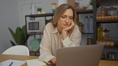 Modern ofis ortamında dizüstü bilgisayarda çalışan, sarı saçlı, odaklanmış, ofis malzemeleri, kapalı mekan bitkileri ve arka planda mutfak malzemeleriyle çevrili bir kadın..
