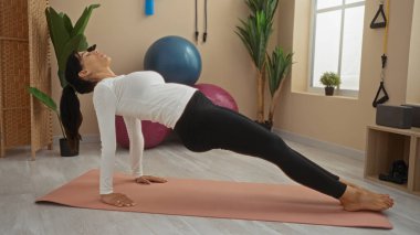 Spor merkezinde yoga yapan bir kadın. Arka planda spor topları ve bitkiler var.