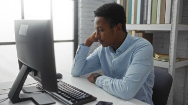 Masaüstü bilgisayarı ve arka plandaki raflarla modern ofis içinde masa başında çalışan mavi gömlekli genç Afrikalı bir adam profesyonel çalışma ortamını gösteriyor.