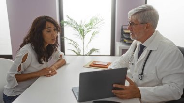Kadın klinikte dizüstü bilgisayarla erkek doktorla konuşuyor, sağlık hizmetlerini tartışıyor; kapalı alanda yapılan toplantı profesyonel tıbbi danışmanlık ortamını canlı bir şekilde yakalıyor.